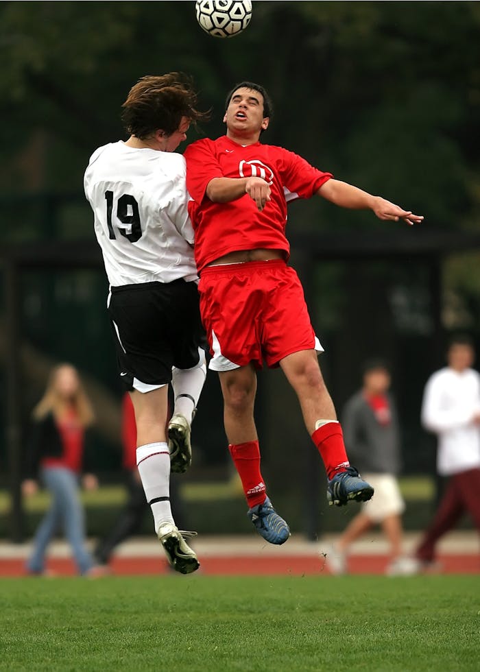 service-04 Two soccer players in mid-air challenge on a green field during a game.